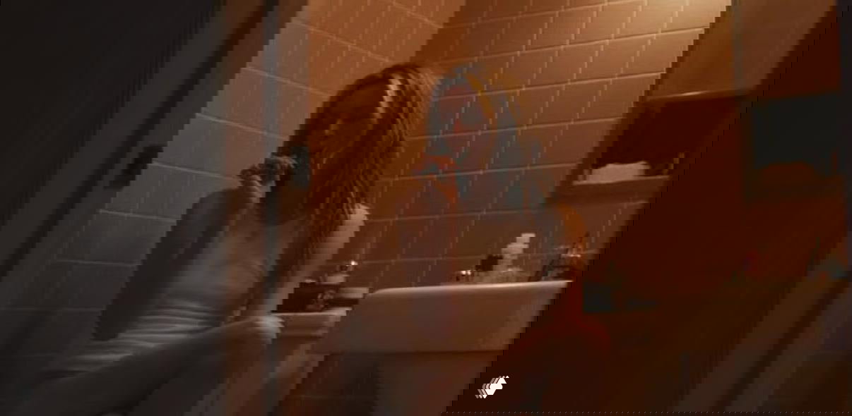 Кадр из сериала: женщина в белой майке сидит в ванной комнате на унитазе, приглушённый свет и интимная, но не откровенная атмосфера сцены.