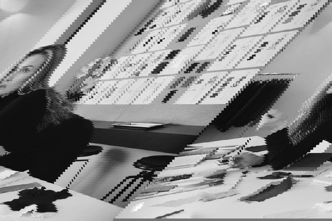 Чёрно‑белый кадр: персонаж в студии дизайна среди образцов и эскизов, минимализм в одежде и рабочая мозаика текстур на заднем плане.