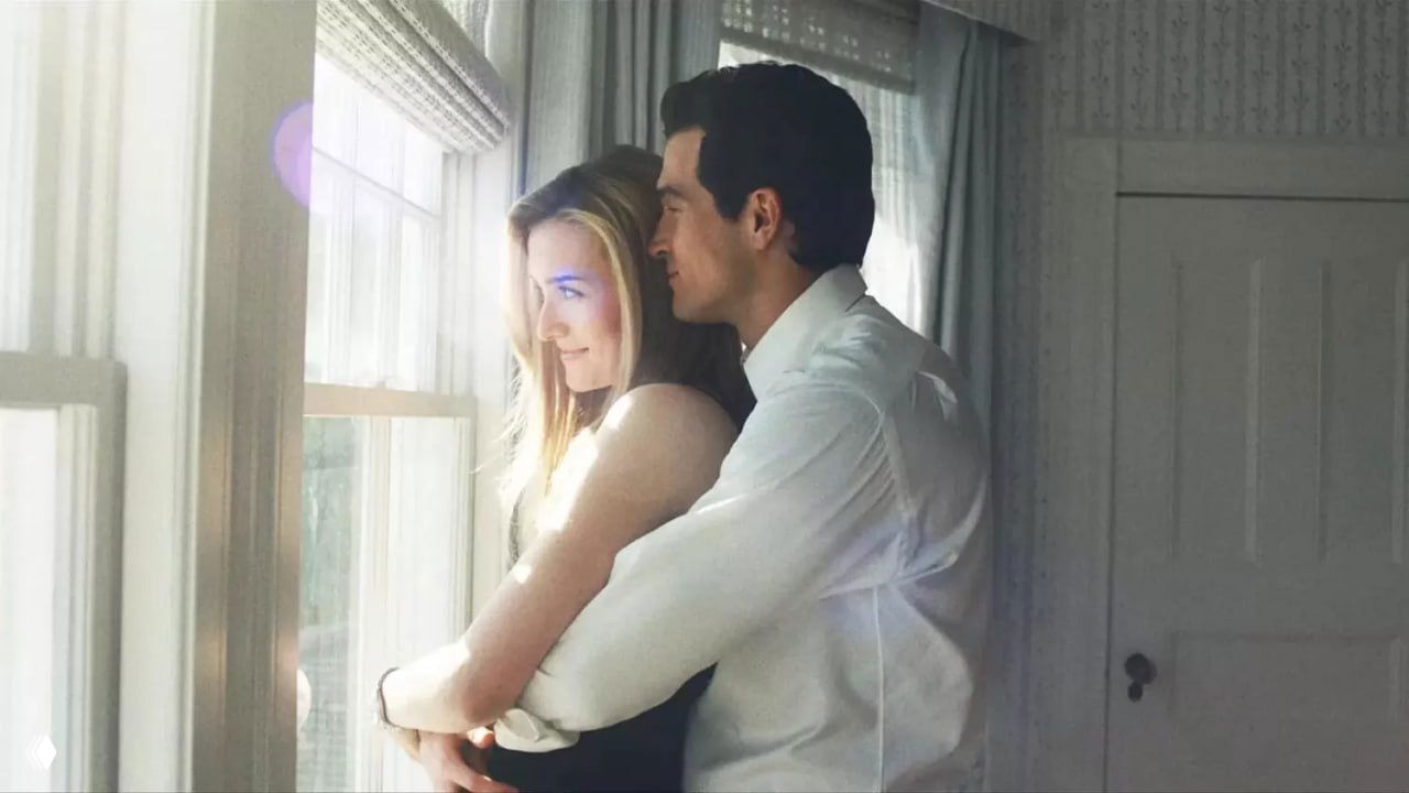 Романтическая сцена у окна: мужчина обнимает женщину, мягкий дневной свет проникает в интерьер, кадр передаёт тихую близость и домашнюю теплоту.