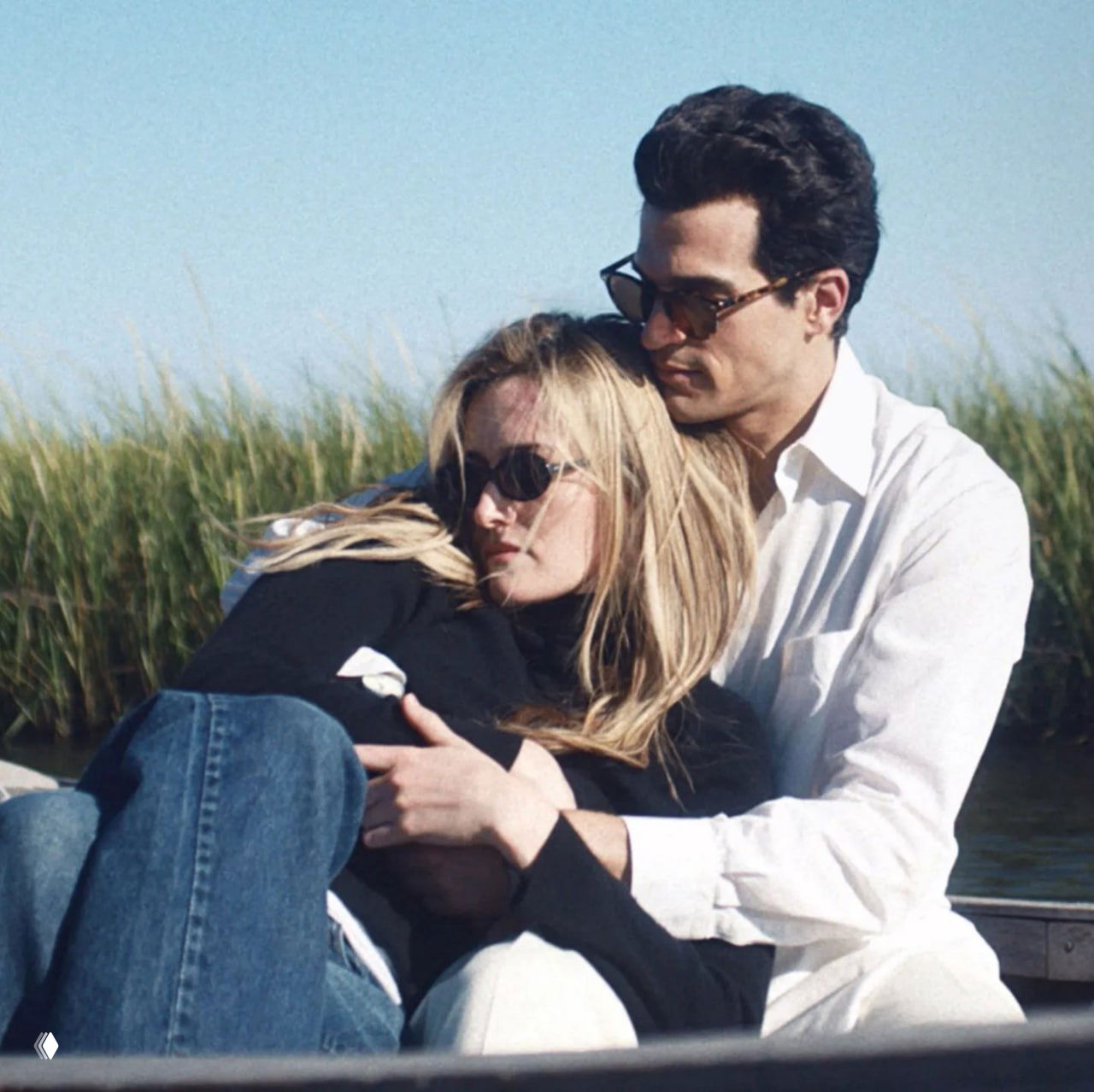 Пара на лодке: мужчина и женщина в солнцезащитных очках обнимаются на фоне тростника и воды, кадр наполнен летней ностальгией и тихой меланхолией.