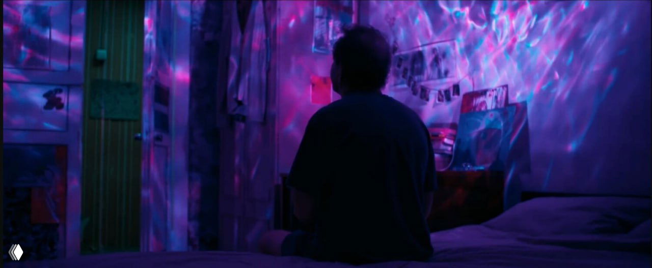 Кадр с цветными световыми проекциями: герой сидит на кровати в полутемной комнате, созерцательная и интимная атмосфера.