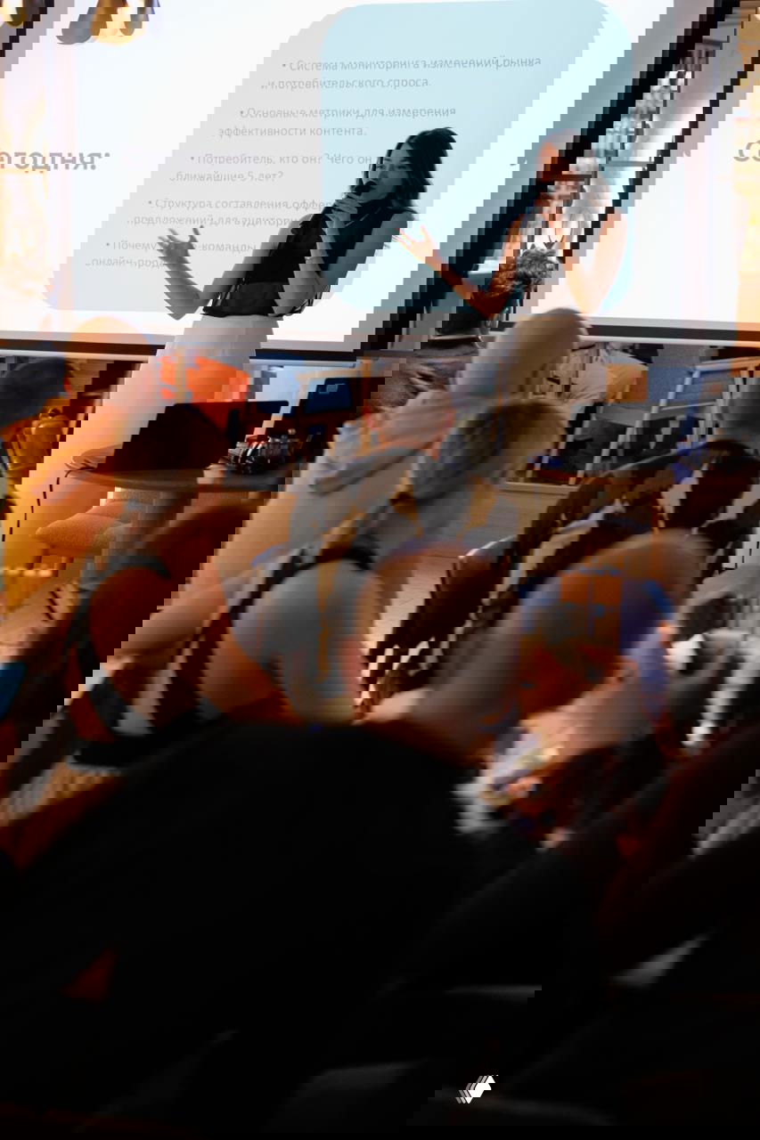 Александра на сцене у проектора, показывает слайды по теме офферов; зрители в первых рядах внимательно следят и записывают ключевые мысли.