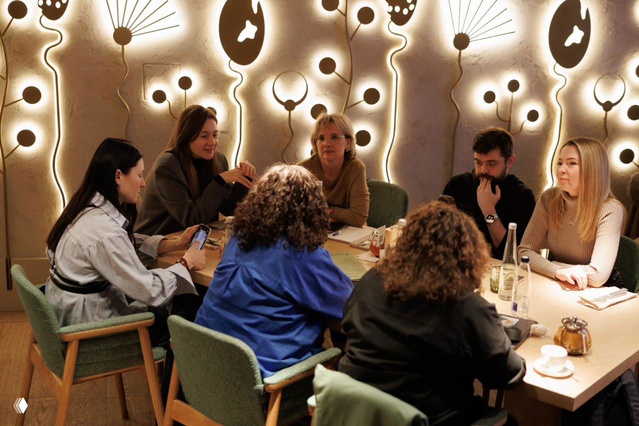 Группа участников за круглым столом обсуждает задания и делится инсайтами; на столе чашки, бутылки воды и блокноты для заметок.