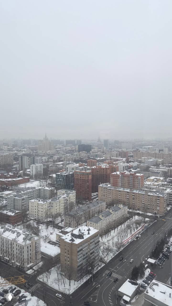 Вид на зимний город из окна: многоэтажные дома, заснеженные улицы и магистраль под серым небом, снимок сверху.