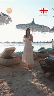 Женщина в платье стоит на песчаном пляже у шезлонгов и зонтов Café del Mar в Шекветили — вид пляжного клуба и летняя атмосфера на побережье.