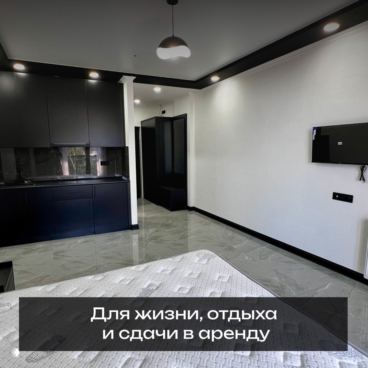 Интерьер готовой студии с современным ремонтом: кровать, чёрная встроенная кухня, глянцевый мраморный пол и потолочные светильники.