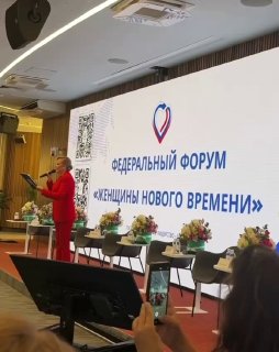 Выступление на сцене федерального форума «Женщины нового времени»: докладчица перед панелью, столы с цветами и участники, снимающие на телефоны.