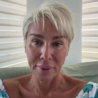 Крупный план женщины со светлыми короткими волосами на фоне светлых штор; нейтральное выражение лица, бытовой интерьер.