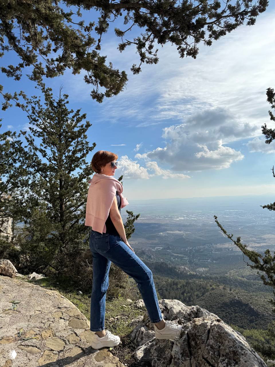 Женщина в походной одежде на вершине утёса смотрит на панораму долины и моря — пейзаж Северного Кипра, момент отдыха и вдохновения.