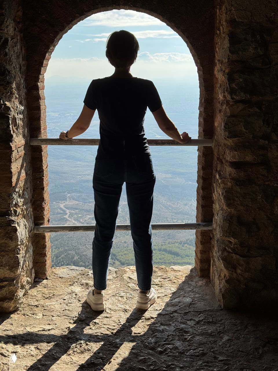 Силуэт женщины, стоящей в арочном окне с видом на горную долину и горизонт — образ созерцания и вдохновения в путешествии.