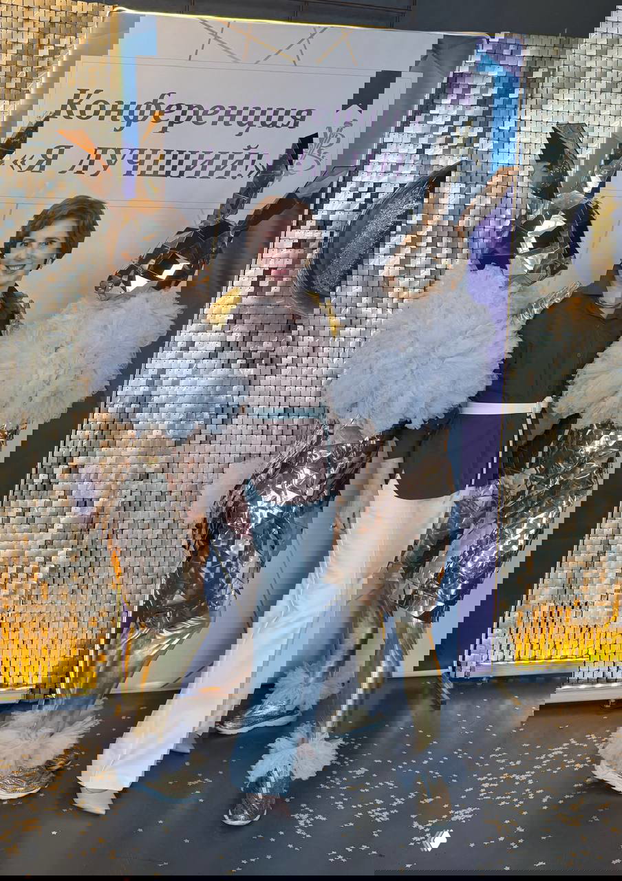 Женщина-спикер позирует между двумя золотистыми ростовыми фигурами у баннера «Конференция «Я ЛУЧШИЙ!»; виден праздничный фон.