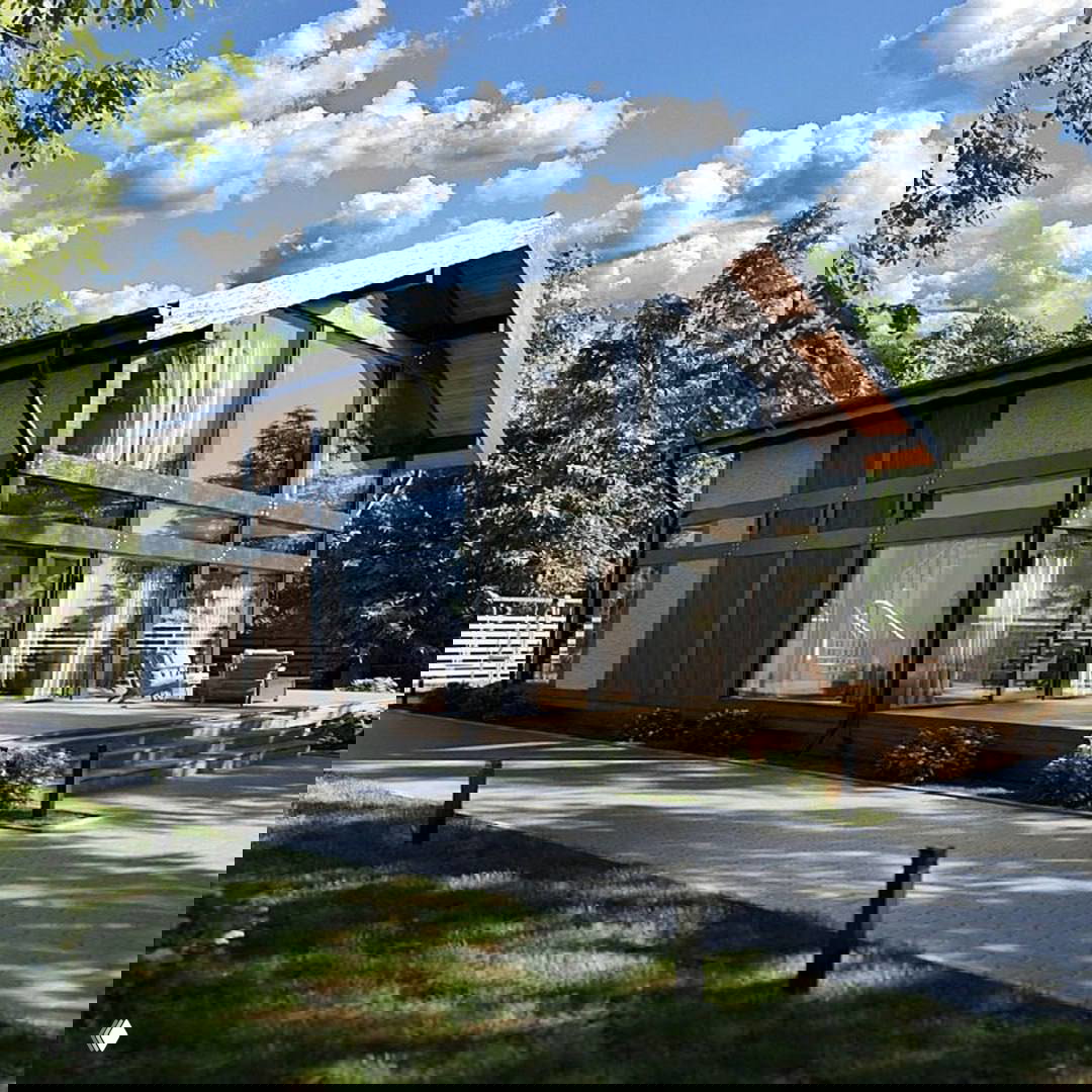 Фасад фахверк‑дома с панорамными окнами и деревянной террасой, вид на сад и окружение из деревьев при дневном свете.