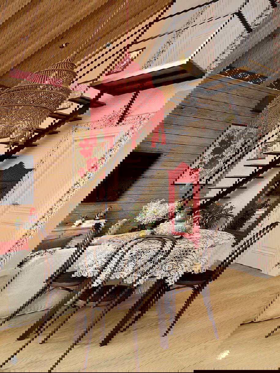 Уютная столовая в фахверк‑доме: деревянные стены, кружевная скатерть на столе и крупная декоративная красная подвесная люстра.