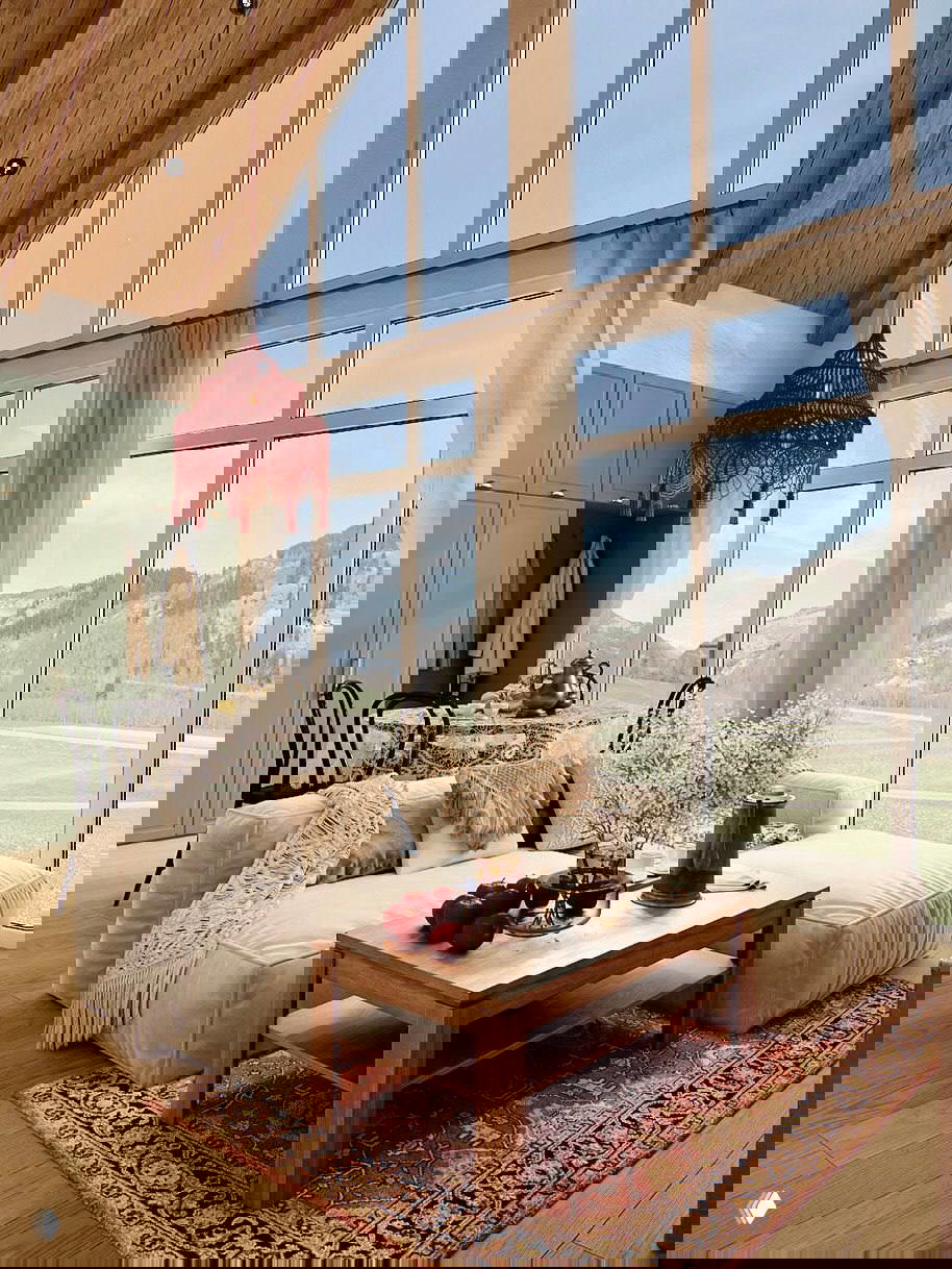 Гостиная с низким диваном, деревянным журнальным столиком и панорамными окнами с видом на поля и горы за участком.