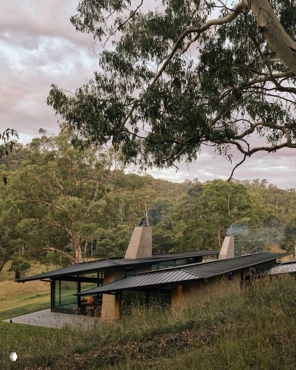 Фрагмент крыши и два скромных дымохода на фоне густых деревьев и холмов, подчёркивающие модульную структуру и интеграцию с рельефом.