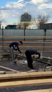 Два строителя на участке частного дома в Астане: укладка лаг на фундамент на фоне забора и бытовок; логотип «Mansion» наложен сверху.