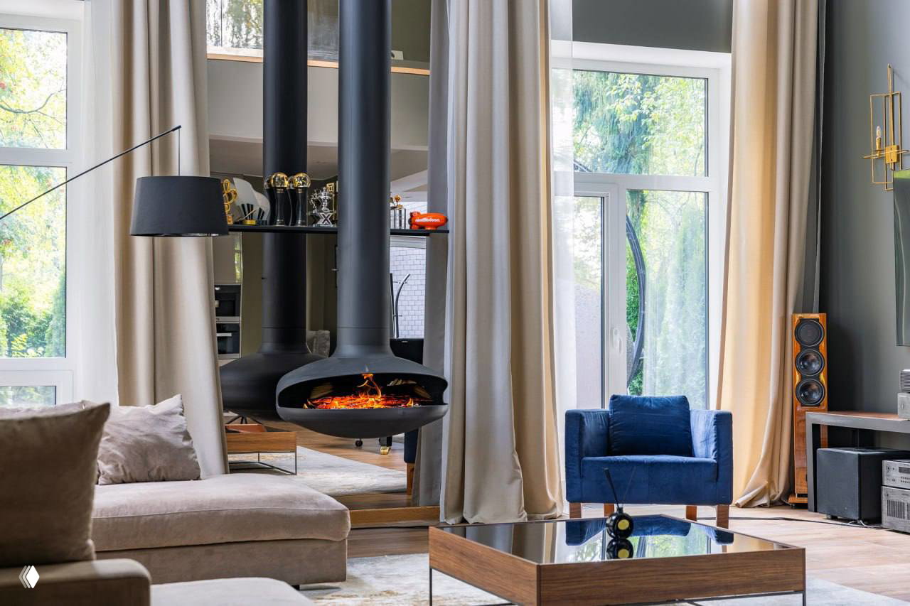 Интерьер гостиной с подвесной каминной печью и высокими окнами в пол, мягкой мебелью и видом на зелёный участок частного дома.