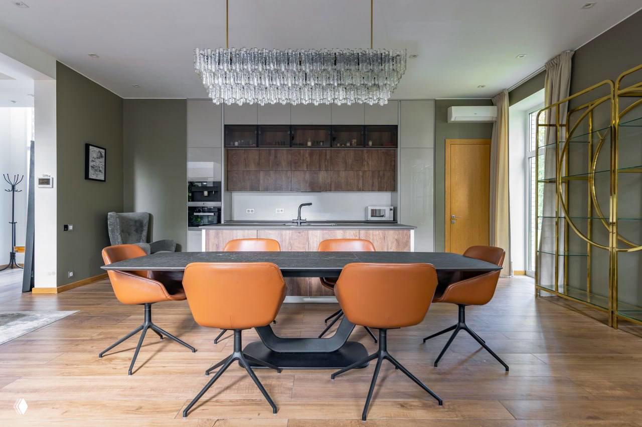 Столовая и кухня в минималистическом стиле: большой обеденный стол, кожаные стулья, встроенная кухня и хрустальная люстра над зоной приёма пищи.