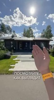 Фасад современного загородного дома при ярком солнечном небе; впереди видна рука с жёлтым браслетом, дорожка и газон.