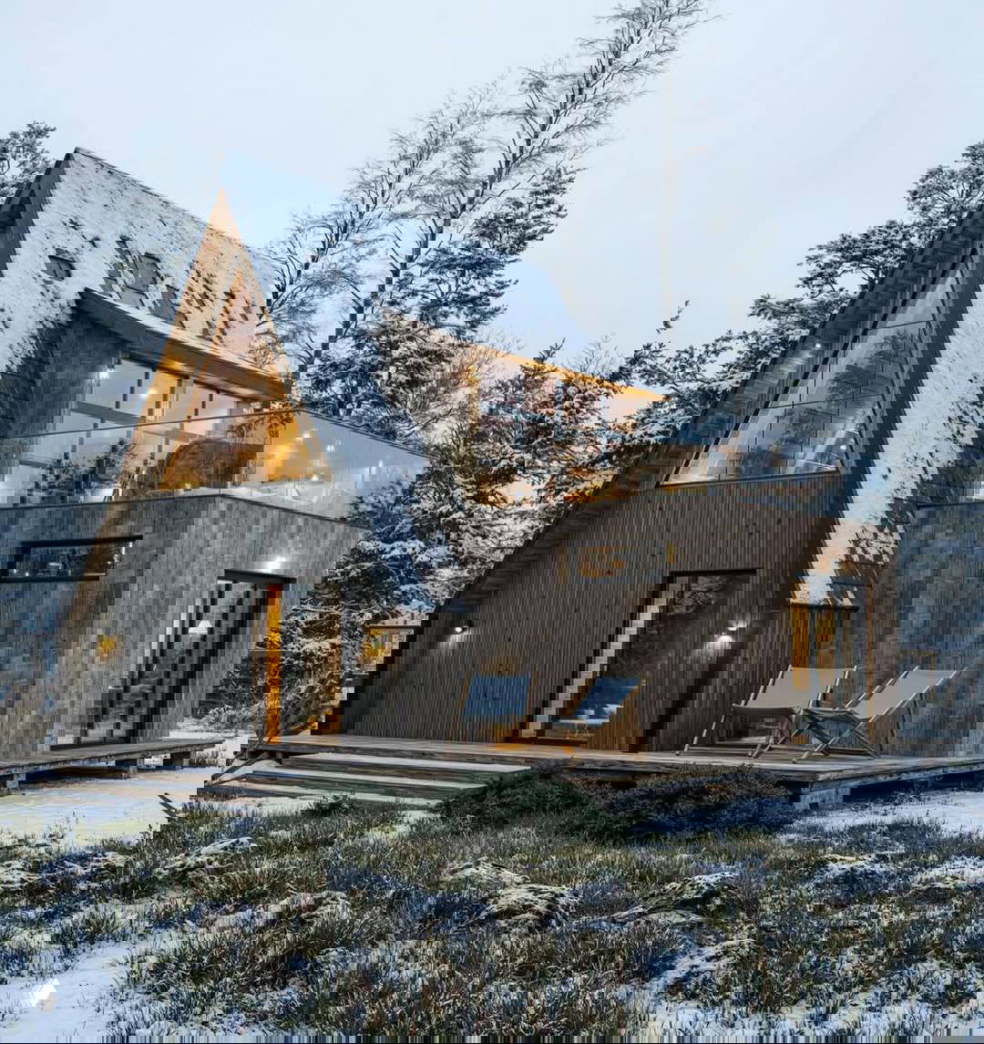 Вид сбоку на деревянный дом с террасой и плоской крышей, заснеженной мансардой и стеклянными ограждениями, окружённый соснами.