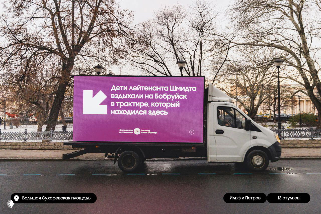 Белый грузовик с большой пурпурной LED-панелью; на экране фрагмент о трактире из «12 стульев», на фоне деревьев и ограждения городского парка.