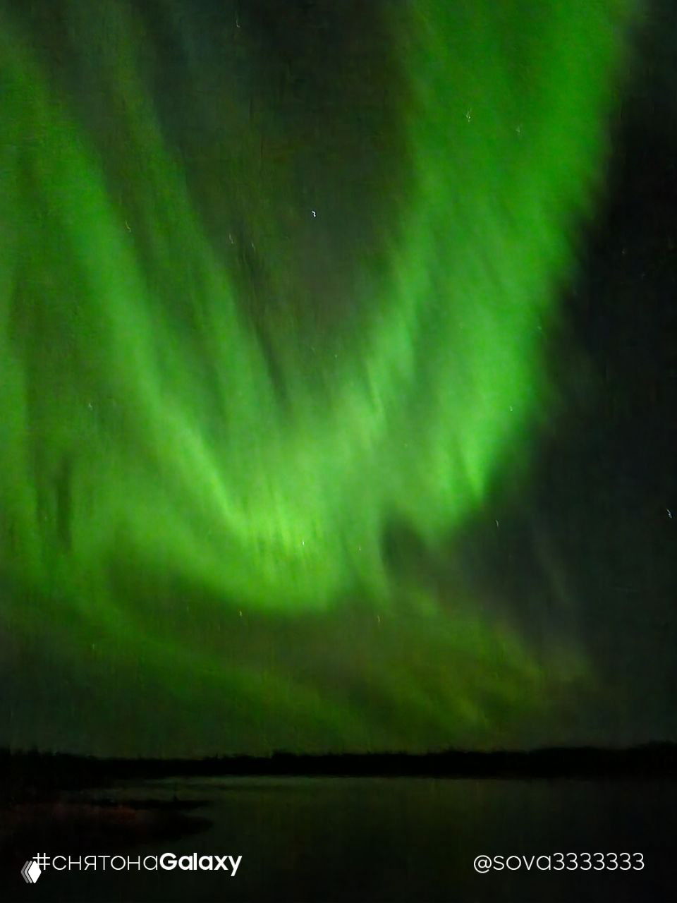 Интенсивные зелёные завихрения северного сияния отражаются в тёмной воде озера на переднем плане, звёзды видны на заднем плане, знак «#снятонаGalaxy» в углу.