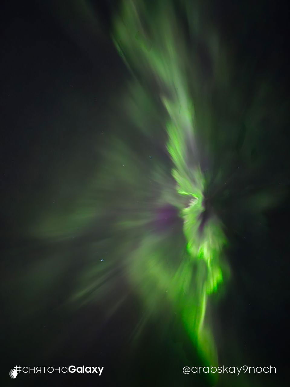 Вертикально ориентированное изображение: динамичные зелёные и фиолетовые потоки полярного сияния, центральная яркая зона и подпись «#снятонаGalaxy» в нижней части кадра.