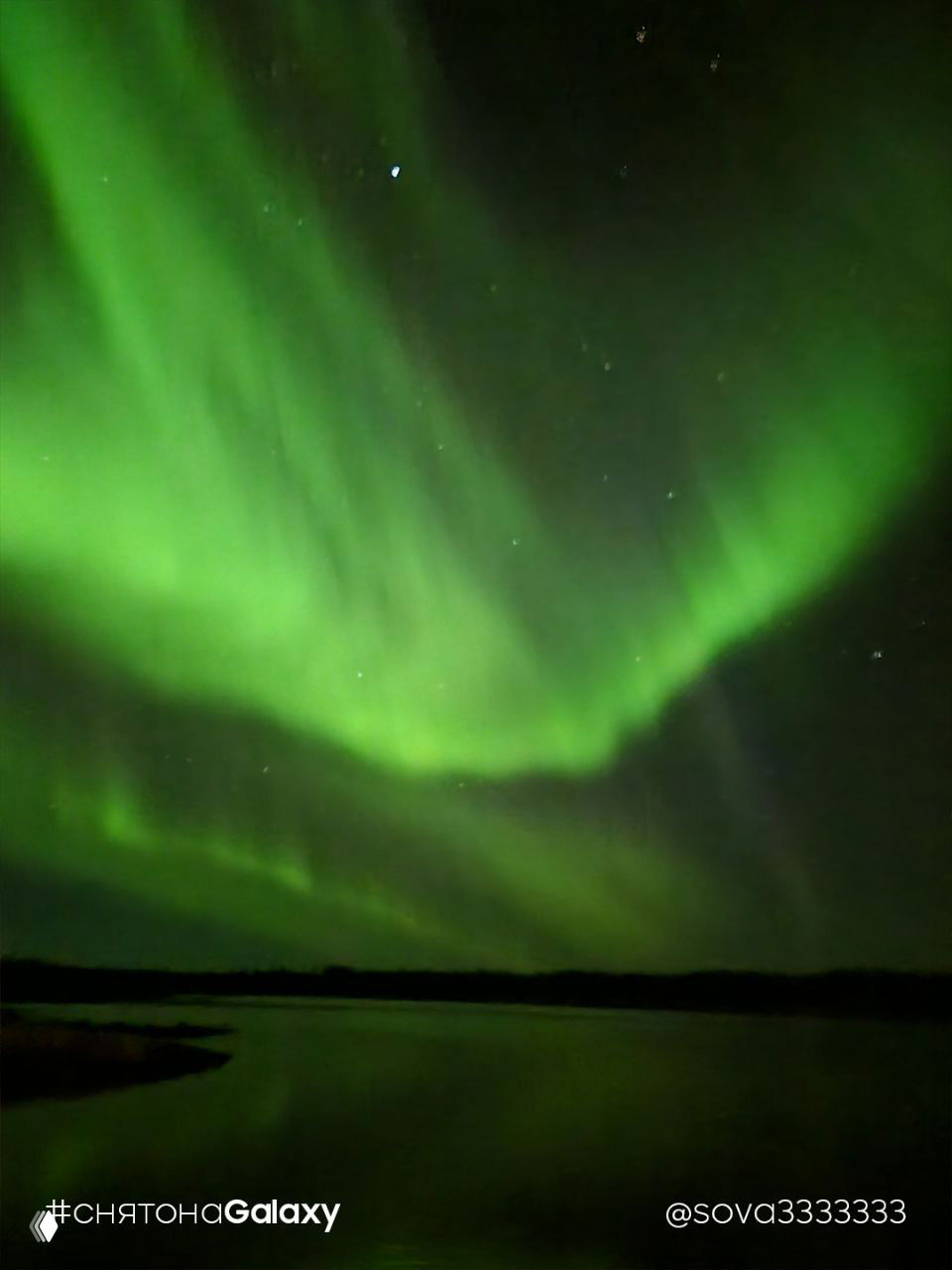 Широкое зелёное полярное сияние над тёмным пейзажем с отражением на воде, мягкие переливы и видны отдельные звёзды, watermark «#снятонаGalaxy» внизу.
