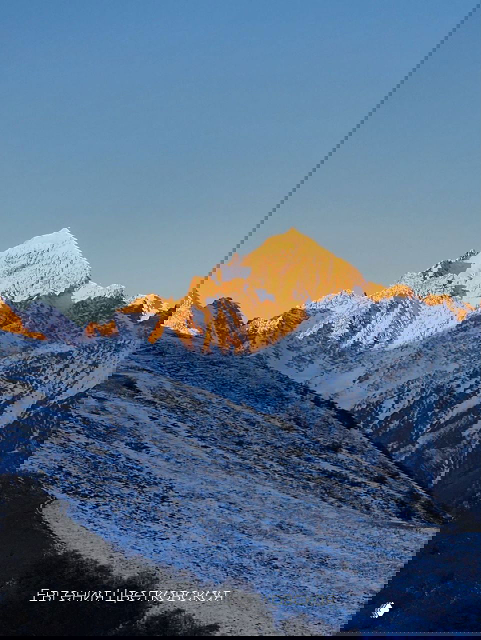 Снежная гора под чистым небом: золотой свет рассвета подчёркивает рельеф склонов и снежные гребни; заметен водяной знак автора снимка.
