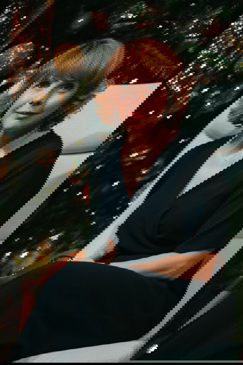 Портрет женщины, сидящей в кресле на фоне праздничной елки; репортажный кадр с литературного вечера в тёплой атмосферной подсветке зала.