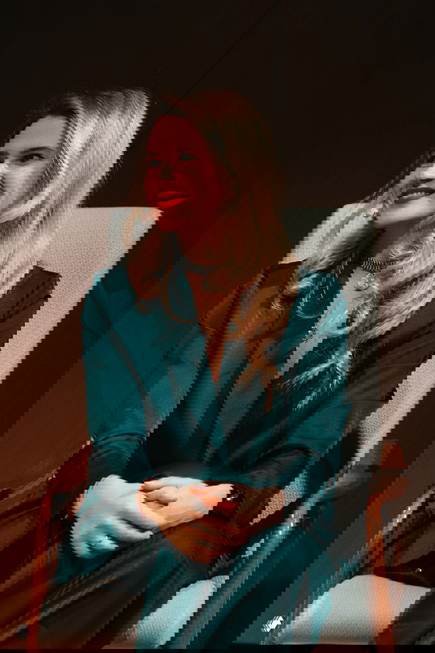 Женщина улыбается, сидя на сцене в кресле; кадр репортажа с мероприятия под рождественские украшения и гирлянды.