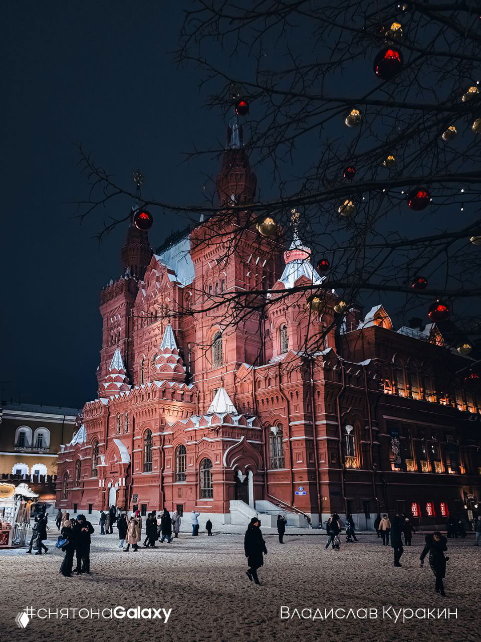 Ночное фото исторического краснокирпичного здания в праздничной подсветке и украшениях, заснеженная площадь и гуляющие люди.