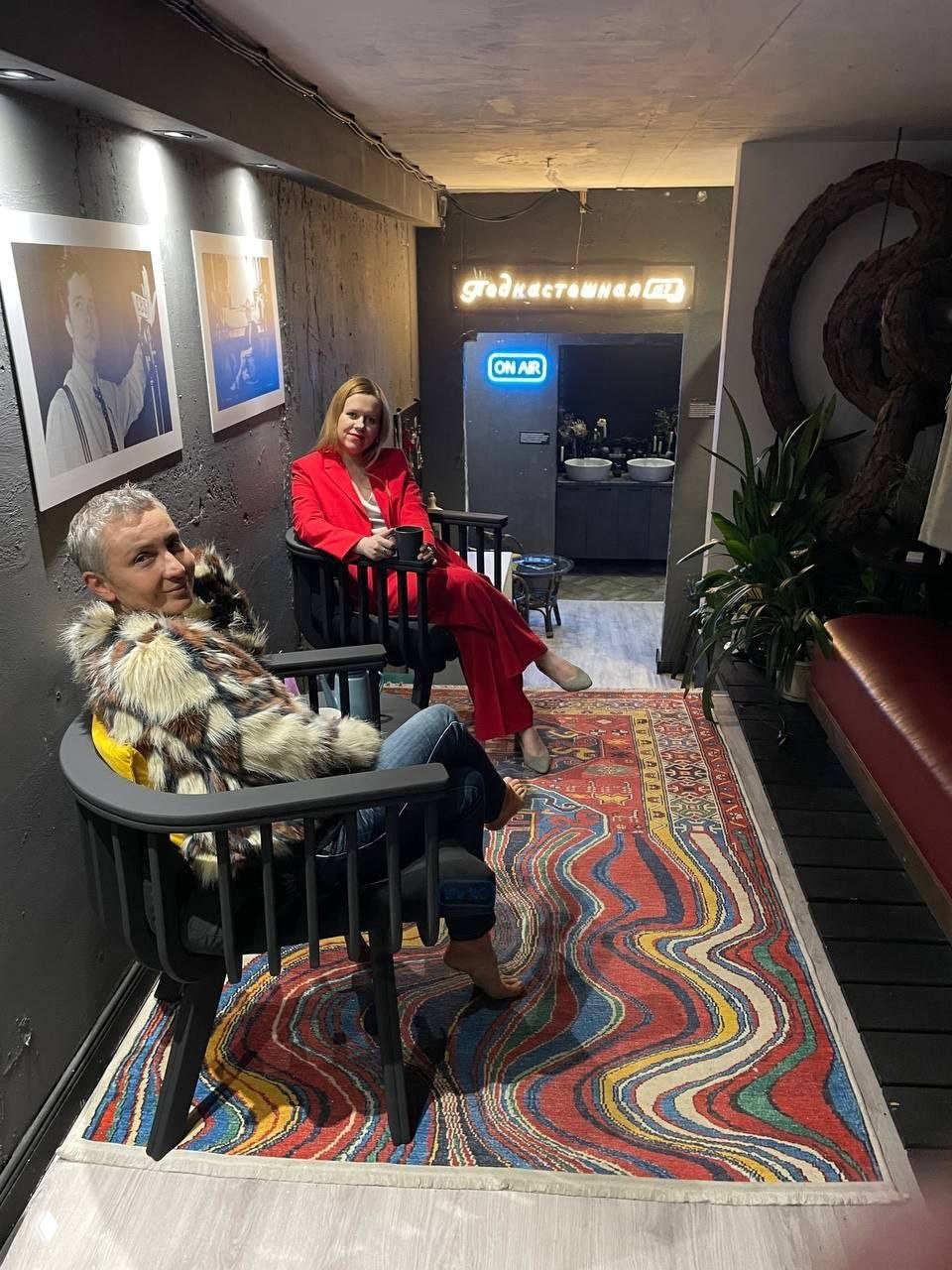 Интерьер Подкастошной № 7: две сидящие гостя на креслах у яркого ковра, неоновая вывеска «ON AIR» и уютная лобби-зона с растениями и фотографиями на стене.