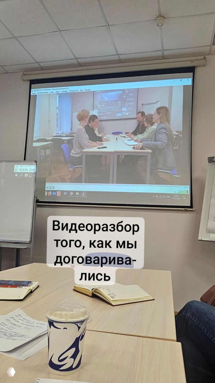 Проектор показывает видеозапись ролевого моделирования переговоров; на столе в переднем плане блокноты и чашка кофе участников тренинга.