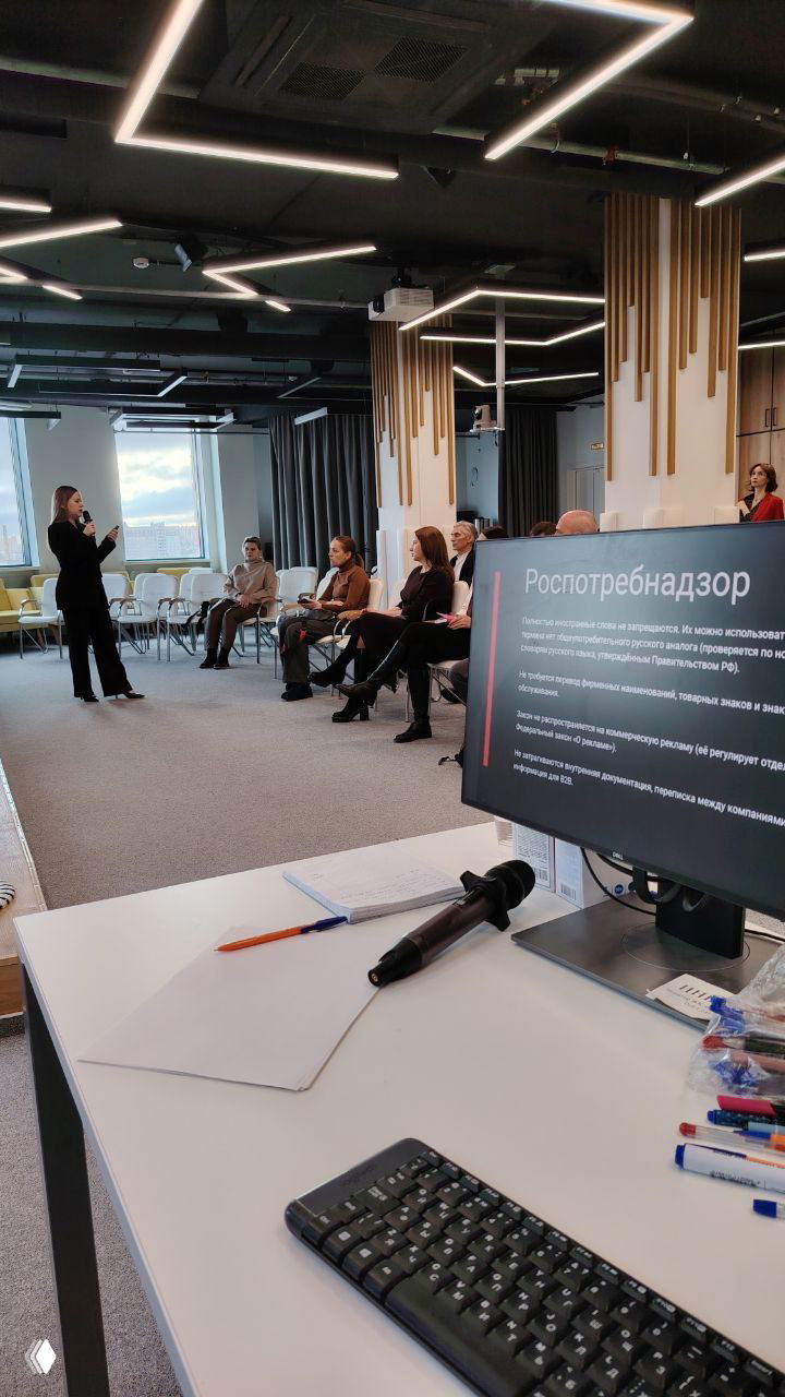 Оратор на сцене перед аудиторией; на переднем плане рабочее место с микрофоном, компьютером и раздаточными материалами для мероприятия.