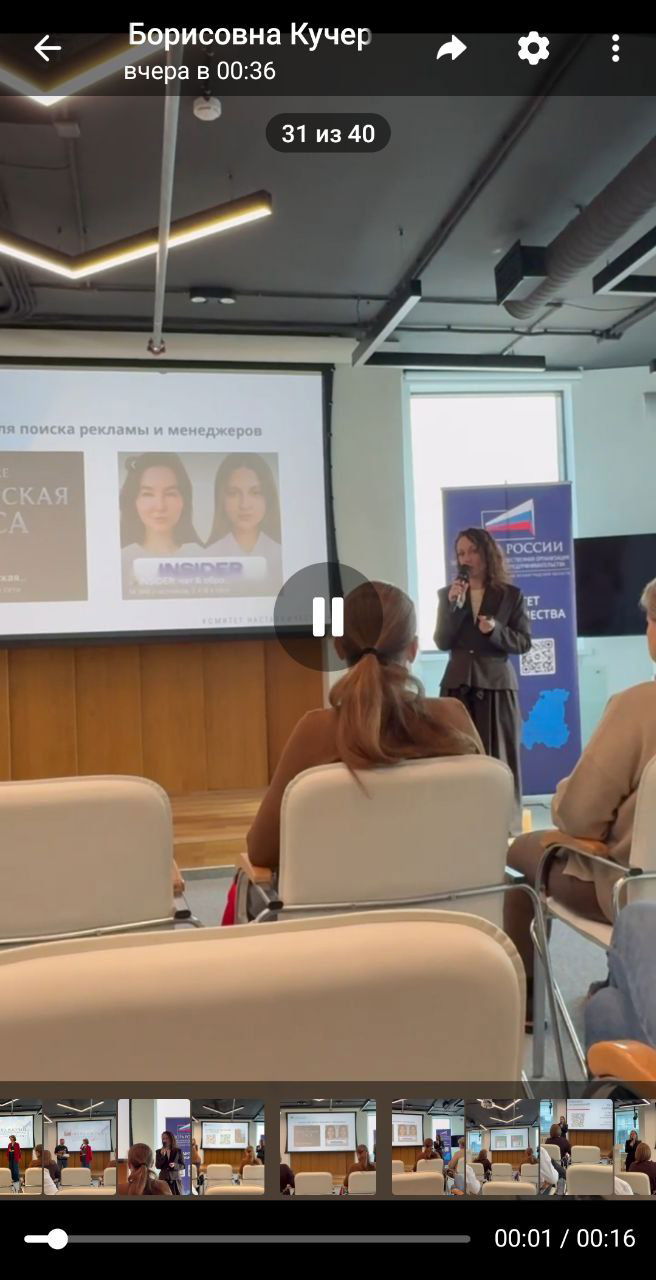 Кадр с места в зале: спикер говорит в микрофон, видна задняя часть головы слушателя и экран с презентацией на фоне.