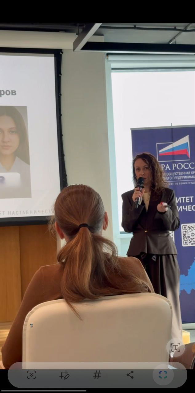 Фотография сессии: женщина выступает перед аудиторией, камера фиксирует кадр с экрана и ряд кресел в зале мероприятия.