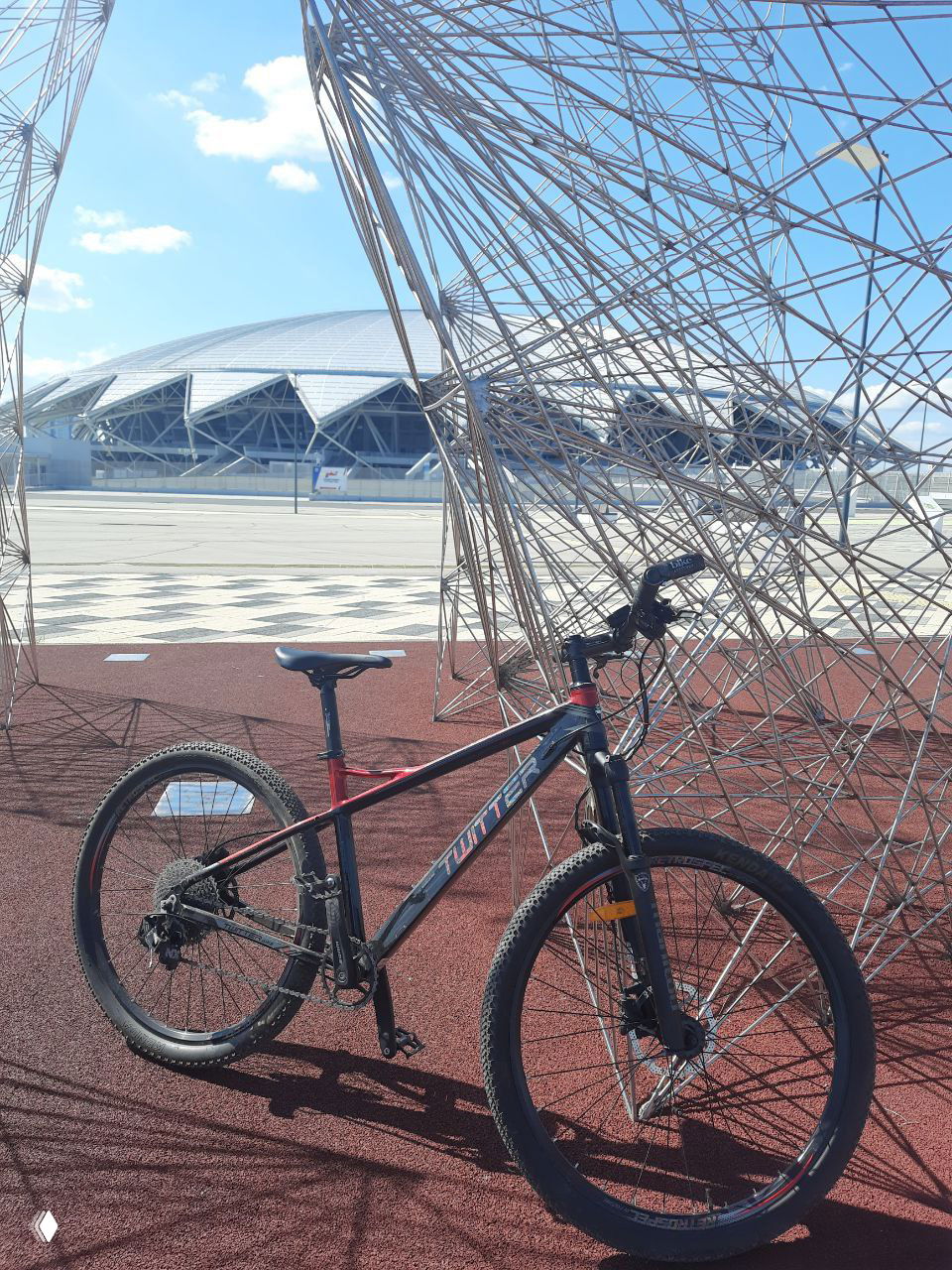 Горный велосипед с надписью бренда на раме установлен рядом с архитектурной инсталляцией; фото подчёркивает дизайн и маркировку велосипеда.