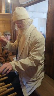 Йозеф Хаазен в белой одежде в кадре внутри карильона, играет на рычагах и педалях — портрет музыканта в работе.