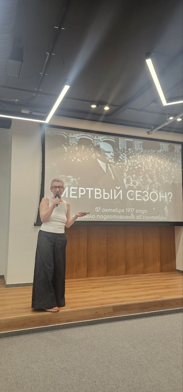 Спикер в светлой майке у экрана с текстом «Мёртвый сезон?», демонстрирует слайд и жестикуляцию во время выступления на мини‑конференции.