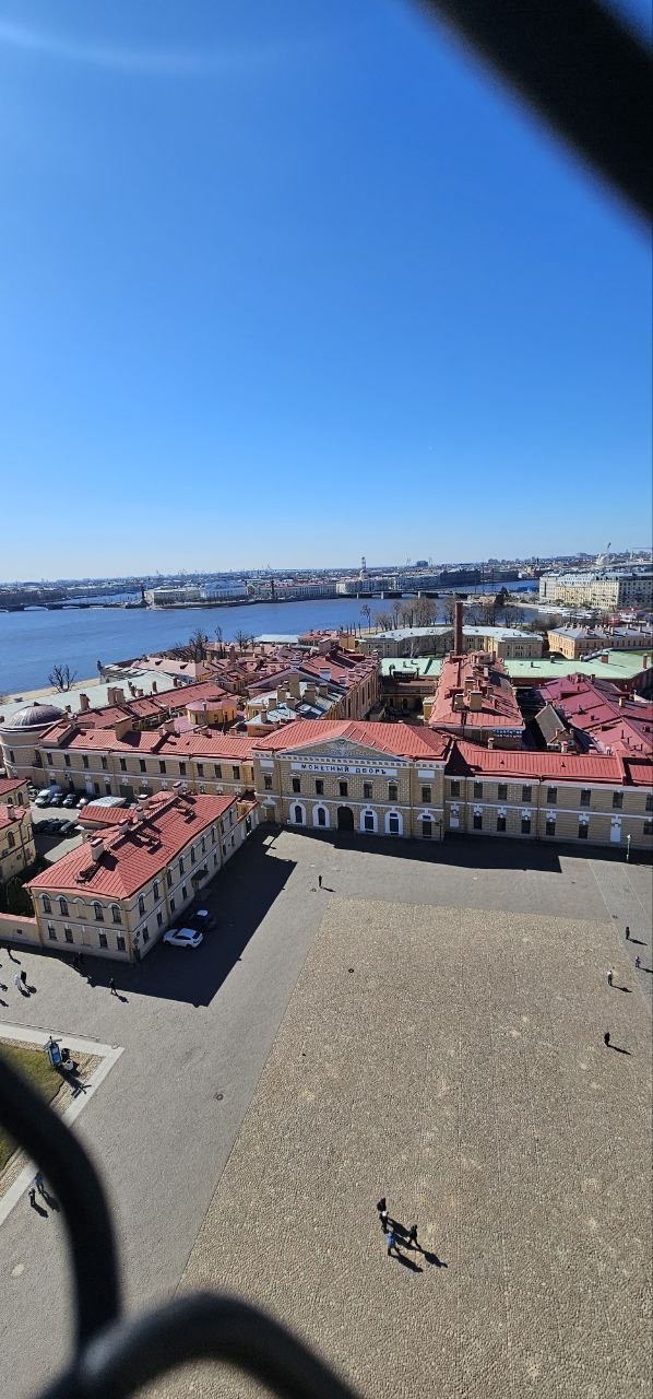 Панорамный вид с высоты 43 метров: двор Петропавловской крепости, крыши зданий и река Нева под ясным голубым небом.