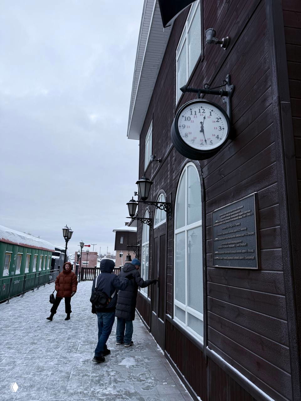 Наружная часть музея техники в зимний день: деревянное здание с настенными часами, макеты вагонов рядом и посетители на заснеженной платформе.