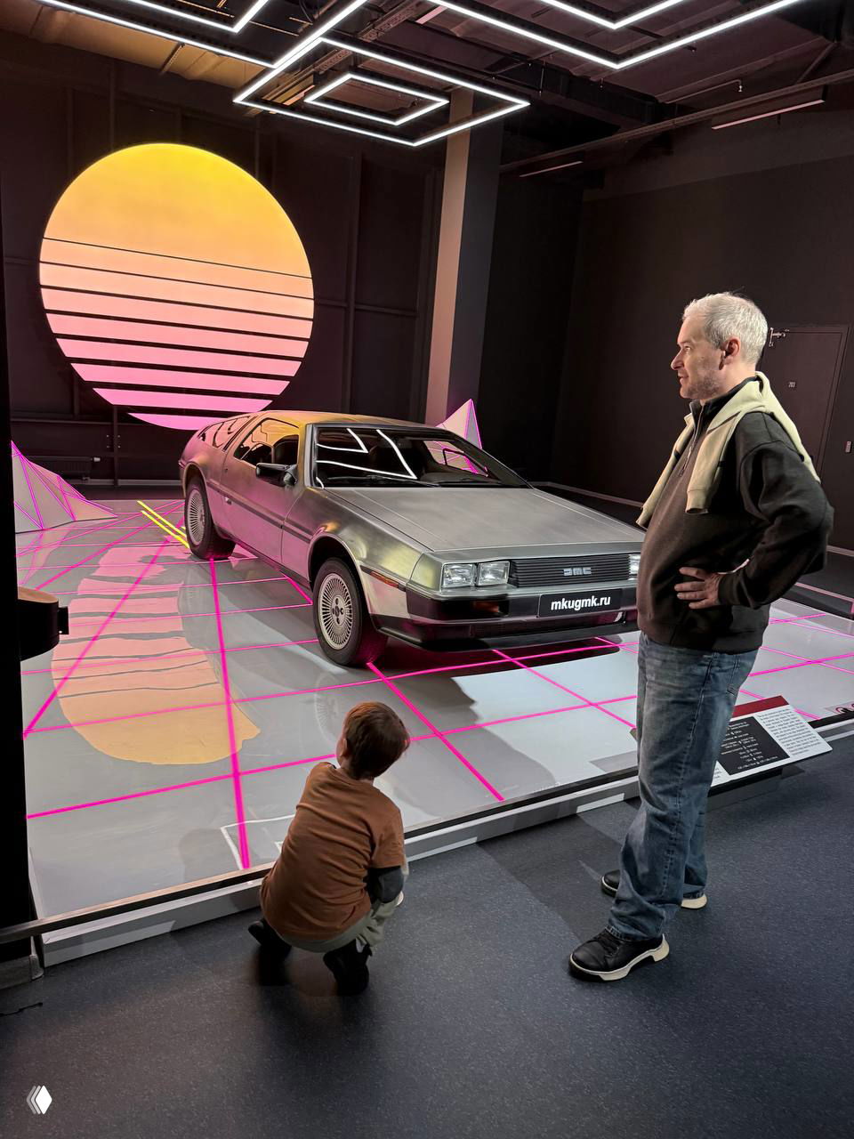 Инсталляция с DeLorean и неоновым фоном: ребёнок и взрослый рассматривают автомобиль в эффектной ретро-футуристической сценографии музея.