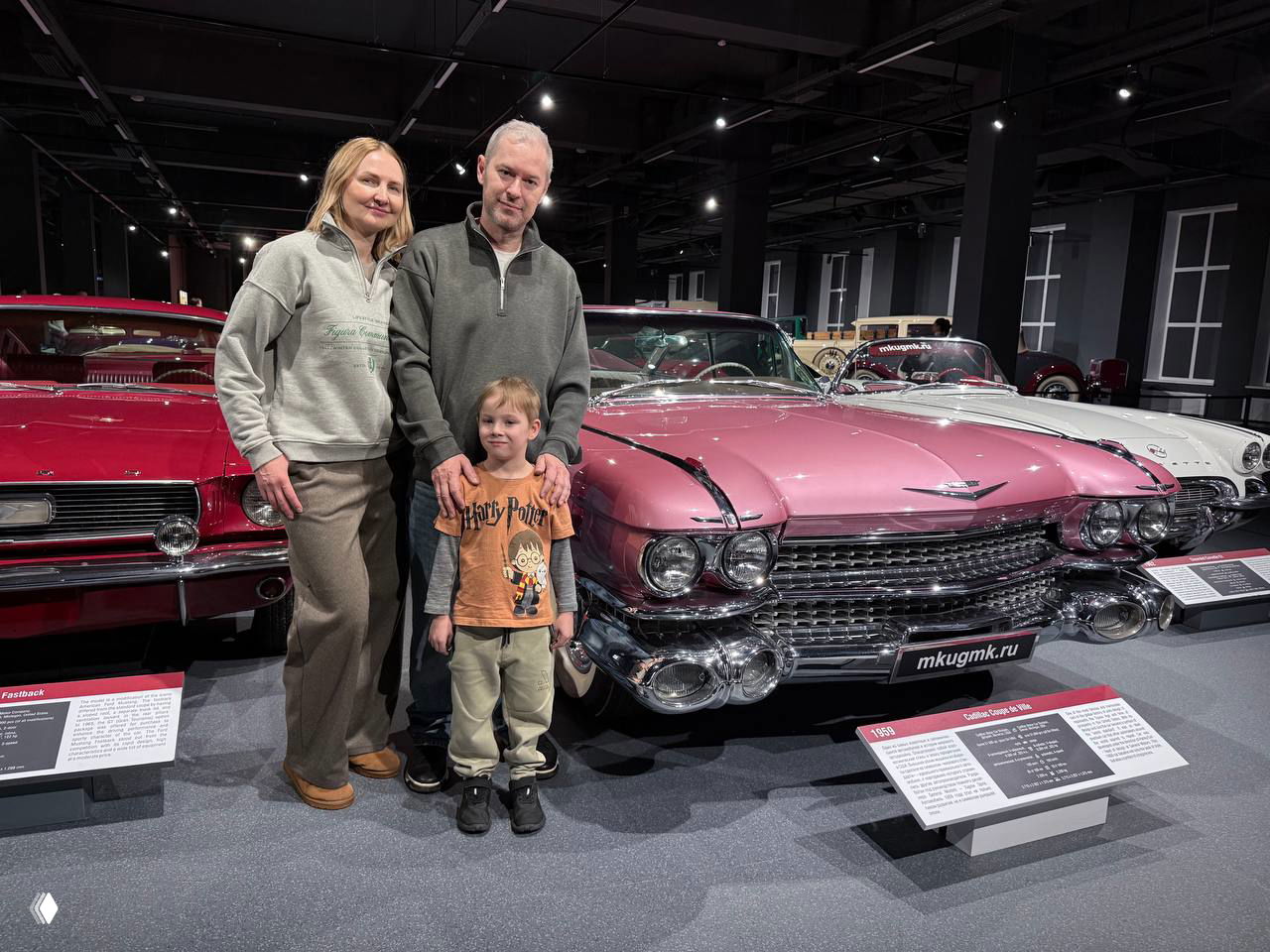 Семейное фото у ряда классических автомобилей: розовый Cadillac и другие ретро-экземпляры, родители и ребёнок на переднем плане.