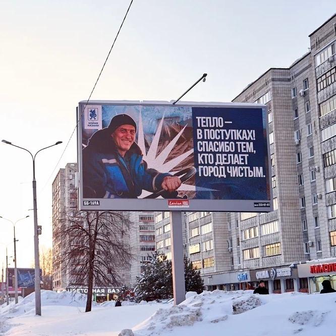 Билборд с улыбающимся работником и надписью благодарности — «спасибо тем, кто делает город чистым» — на фоне домов и сугробов в зимнем городе.