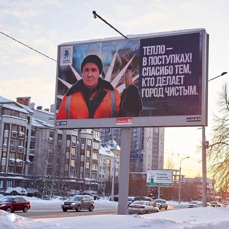 Широкий план улицы с большим билбордом, движением автомобилей и городской застройкой; зимний кадр с рекламой муниципального характера и снегом вдоль дорог.