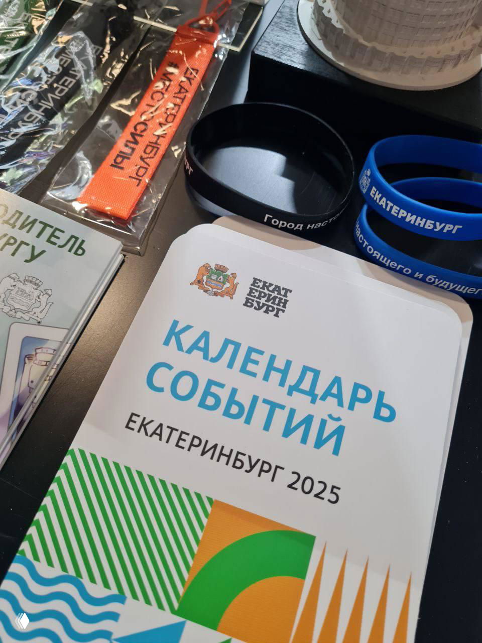 Стол с памятными предметами к юбилею Екатеринбурга: календарь событий 2025, браслеты и брендированные аксессуары с логотипами города.