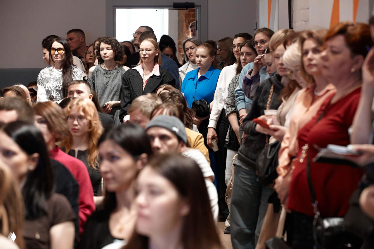 Группа людей стоит и внимательно слушает выступление в коридорной зоне, часть аудитории сфокусирована на спикере и экране.