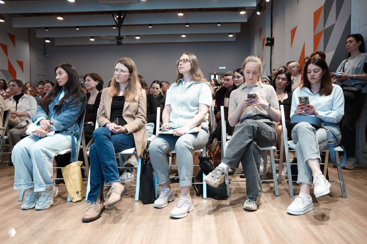 Передний ряд слушателей, некоторые с телефонами и блокнотами, участники внимательно наблюдают за презентацией в зале.