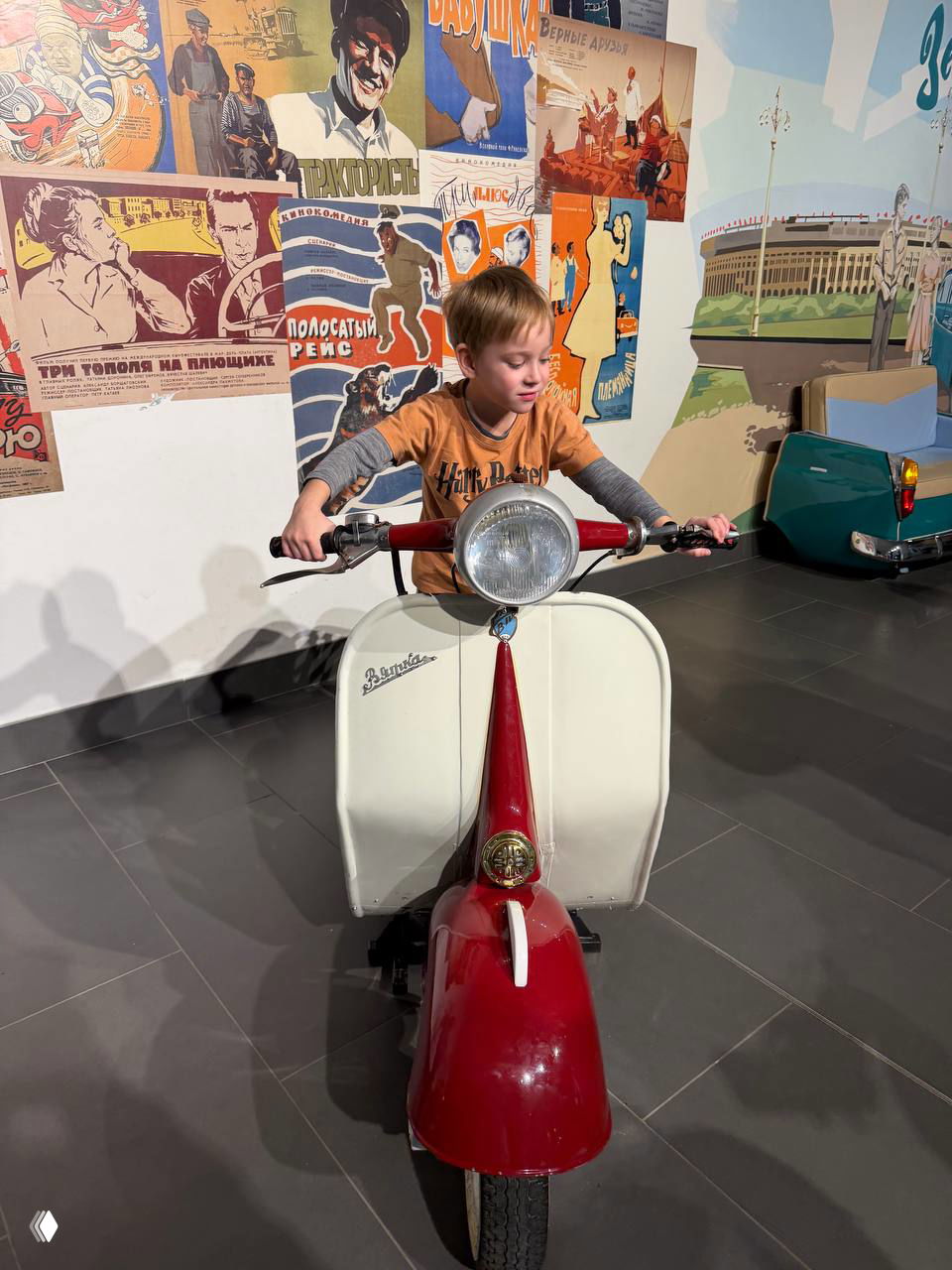 Ребёнок сидит на ретро-скутере внутри выставочного зала, ярко-красный кузов и плакаты советской эпохи на стене за экспонатом.